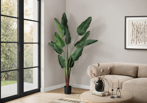 A tall artificial Bird of Paradise plant in a black pot stands in the corner of a living room next to a glass door.