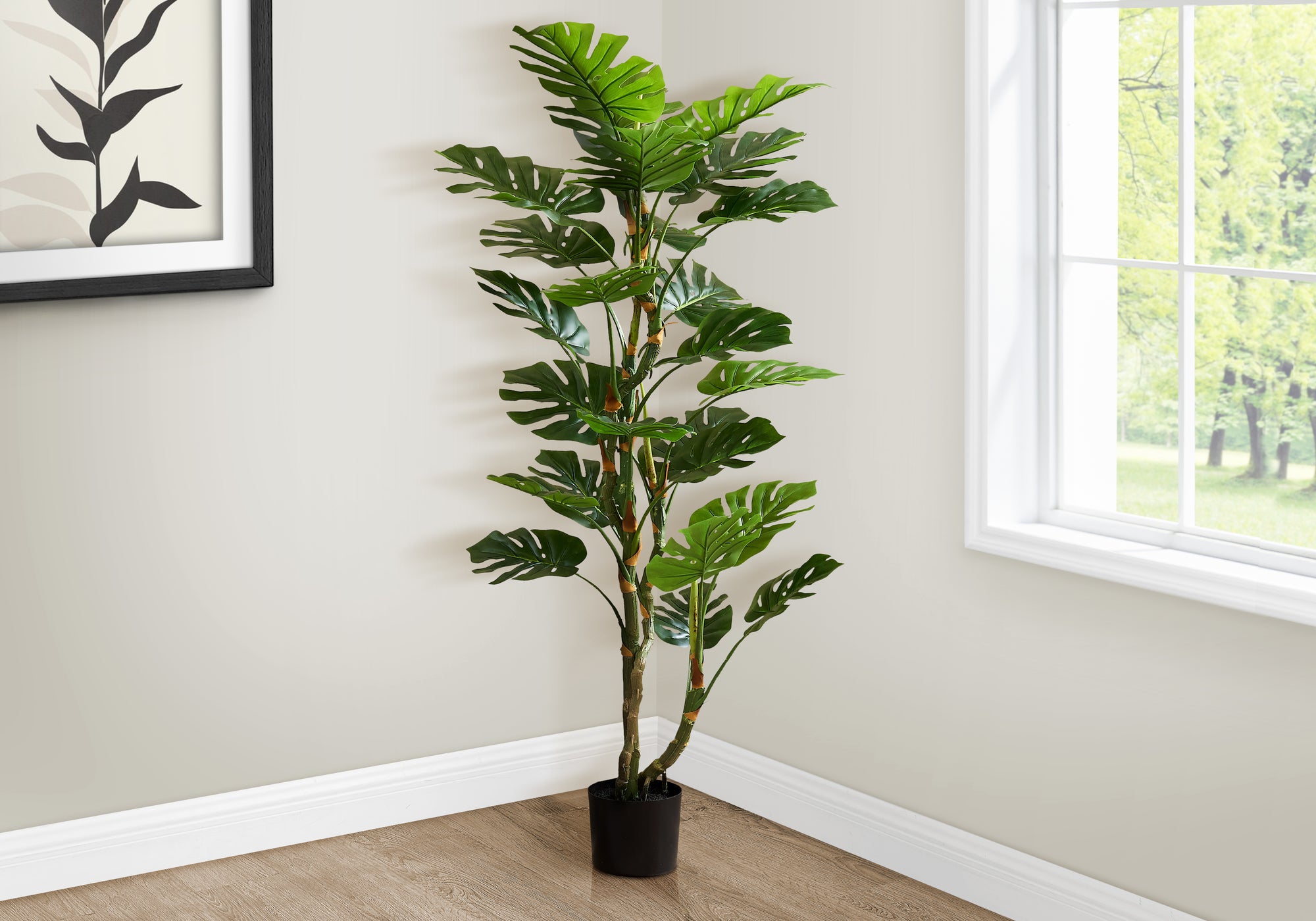 A tall artificial Monstera tree with dual trunks and split leaves standing in the corner of a bright room.