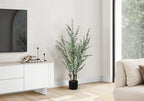 A tall artificial eucalyptus plant in a black pot standing next to a white media console in a bright living room.