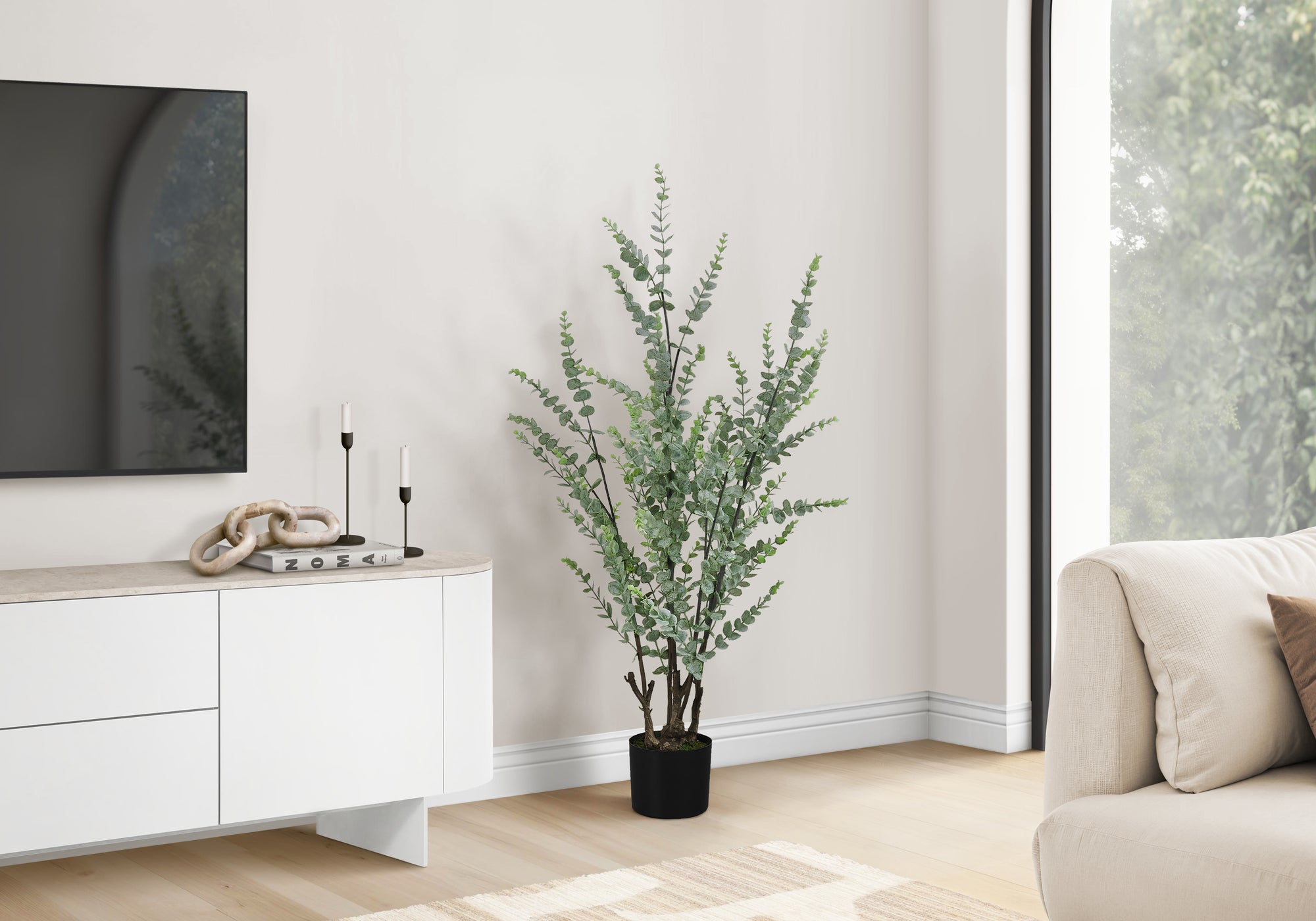 A tall artificial eucalyptus plant in a black pot standing next to a white media console in a bright living room.