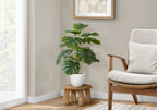 Artificial medium Monstera plant with split green leaves in a white pot sitting on a small wooden stool.