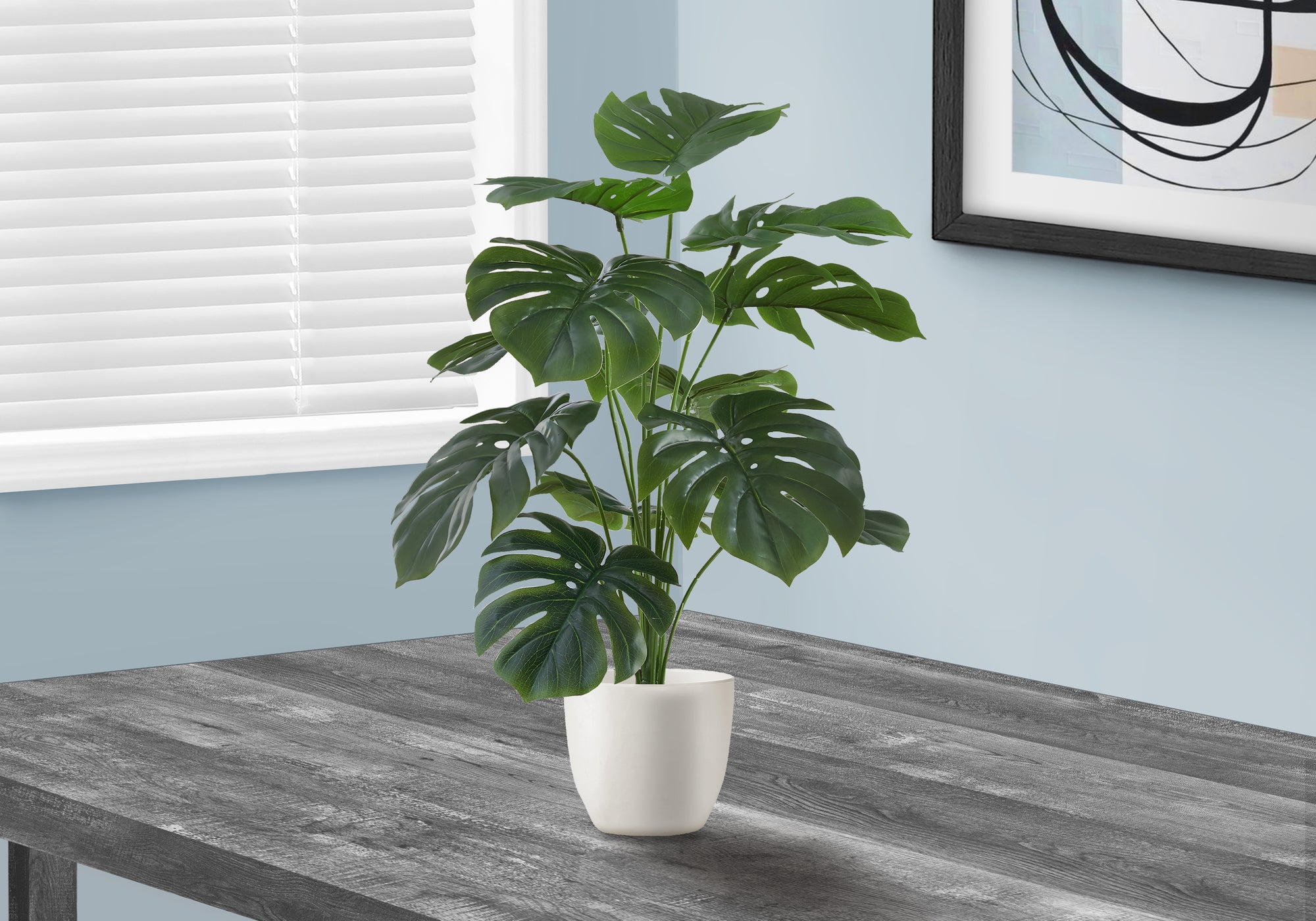 Artificial medium Monstera plant with split green leaves in a white pot, sitting on a dark wooden table.