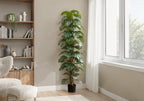 Tall artificial Monstera plant with split leaves climbing a central moss pole in a bright living room corner.